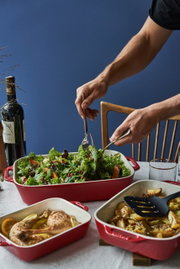 Drie rode ovenschaaltjes met verschillende gerechten, waaronder een frisse salade, kippenboutjes en gepofte aardappelen, van bovenaf gefotografeerd op een tafel.