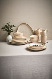 Ein 16-teiliges Kombiservice in Cremefarben auf einem Tisch, bestehend aus Tellern, Schalen und Tassen, mit einer Pflanze im Hintergrund. Perspektive von vorne.