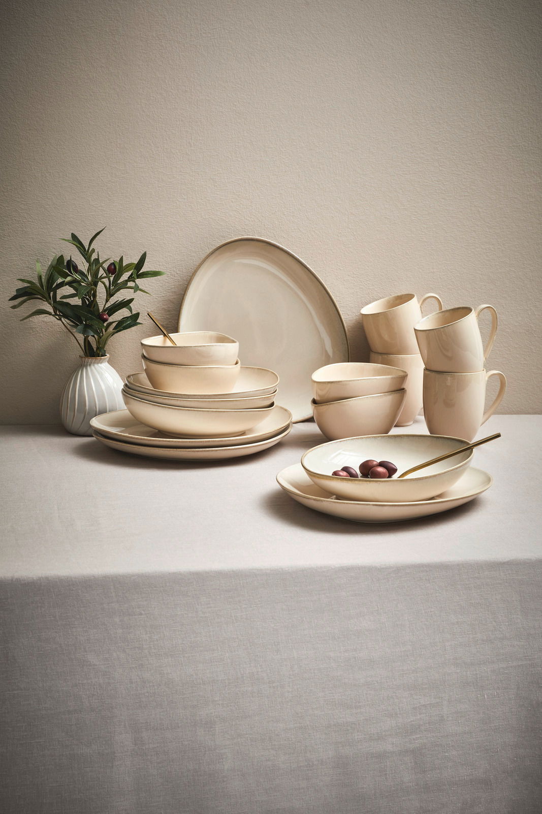 Ein 16-teiliges Kombiservice in Cremefarben auf einem Tisch, bestehend aus Tellern, Schalen und Tassen, mit einer Pflanze im Hintergrund. Perspektive von vorne.