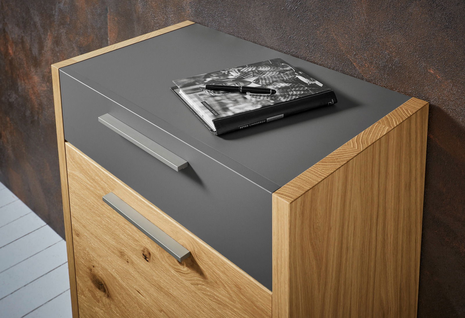 Kommode aus Holz mit grauer Oberfläche, seitliche Perspektive, darauf liegt ein Buch und ein Stift.