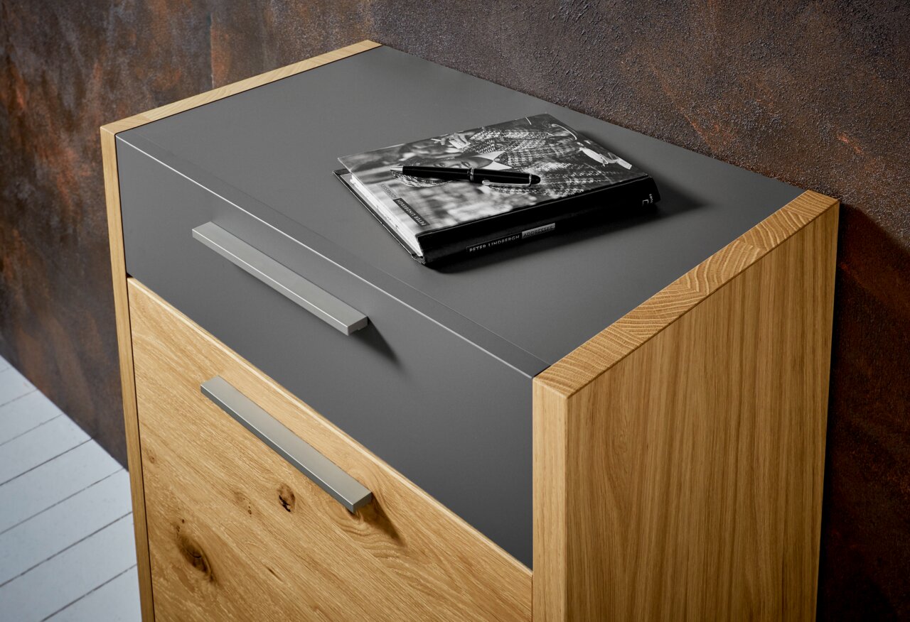 Kommode aus Holz mit grauer Oberfläche, seitliche Perspektive, darauf liegt ein Buch und ein Stift.