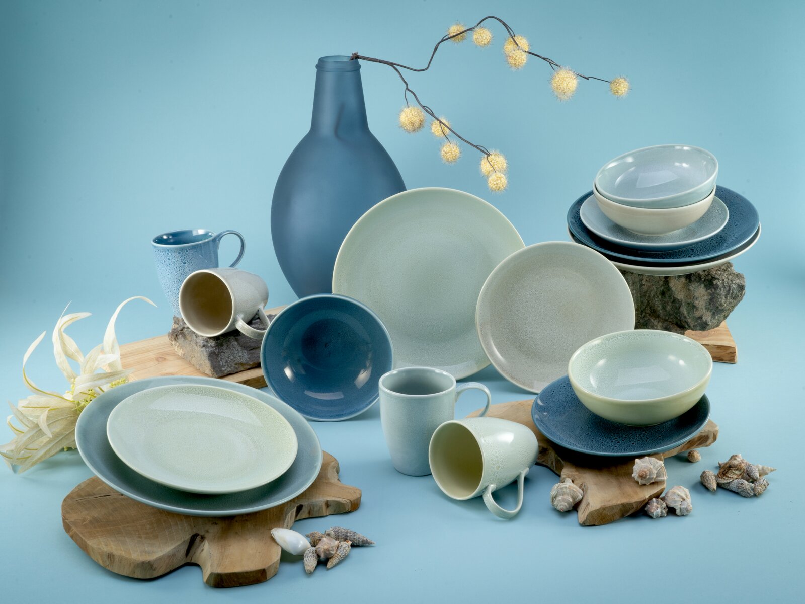 Modernes Keramik-Geschirrset in Blau- und Minttönen mit Tellern, Schalen und Bechern, arrangiert mit großer blauer Vase auf Holzplatten und Steinen vor hellblauem Hintergrund, Perspektive: frontale Studioaufnahme in leichter Aufsicht.