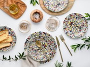 Draufsicht auf bunt gemustertes Geschirr-Set mit Teller und Schale, goldenem Besteck, Glas und Olivenöl auf weißem Hintergrund, dekoriert mit Olivenzweigen und Holzaccessoires