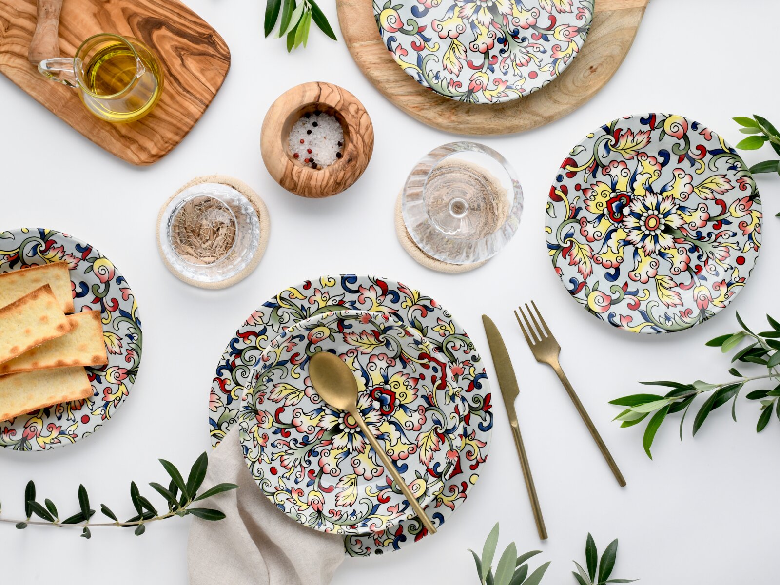 Draufsicht auf bunt gemustertes Geschirr-Set mit Teller und Schale, goldenem Besteck, Glas und Olivenöl auf weißem Hintergrund, dekoriert mit Olivenzweigen und Holzaccessoires