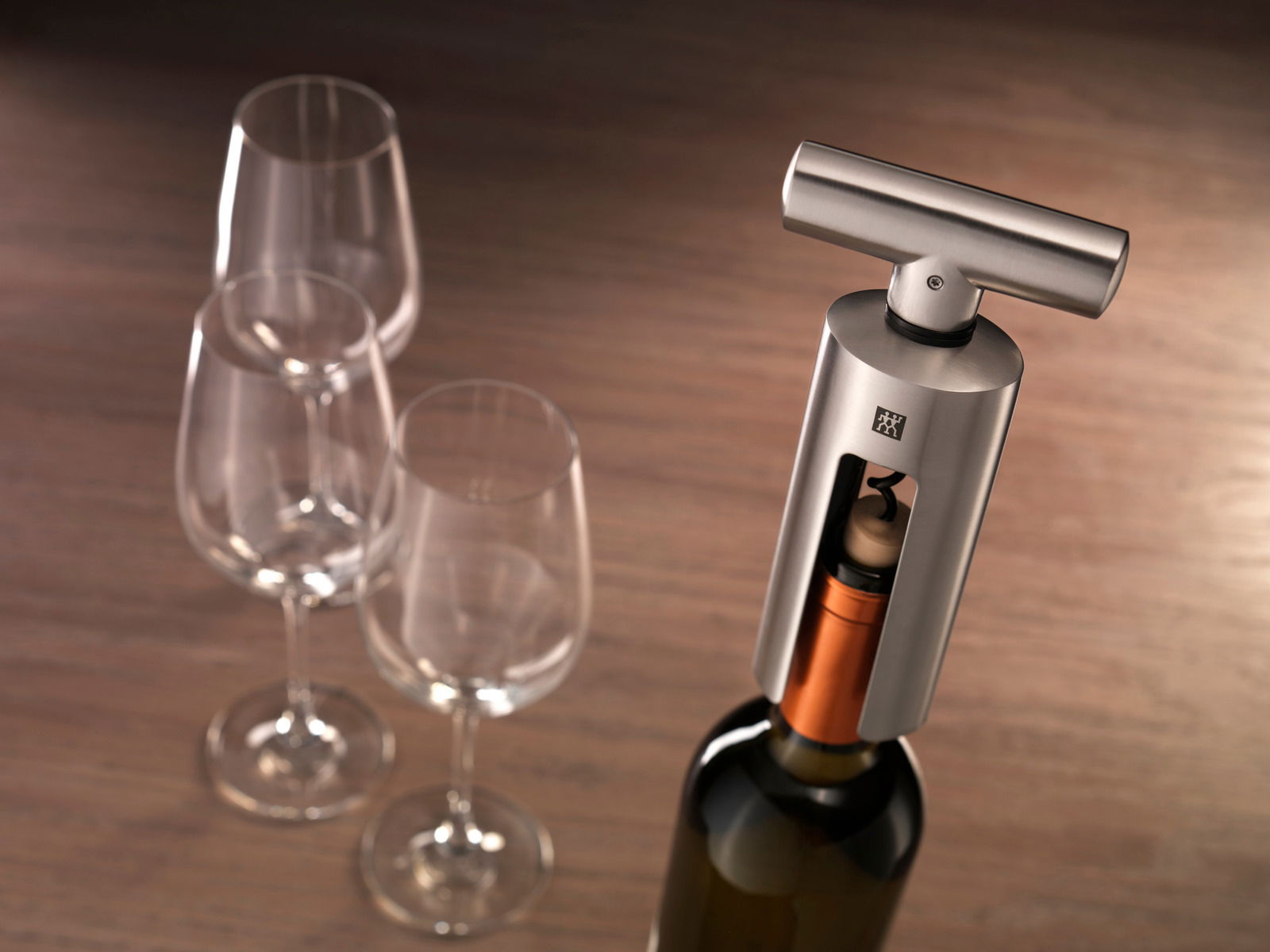 Moderner Korkenzieher aus Edelstahl in Nahaufnahme, neben drei Weingläsern auf einem Holztisch.