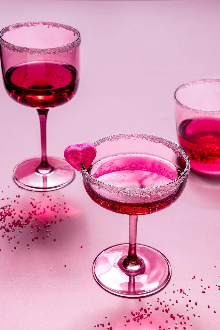 Zwei rosafarbene Weinkelche mit Zuckerrand und Herzdekoration, aus der Vogelperspektive fotografiert.