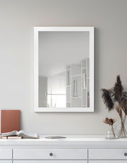 Rechteckiger Spiegel mit weißem Rahmen, an einer grauen Wand hängend, mit einer Kommode im Vordergrund. Perspektive: Frontalansicht.