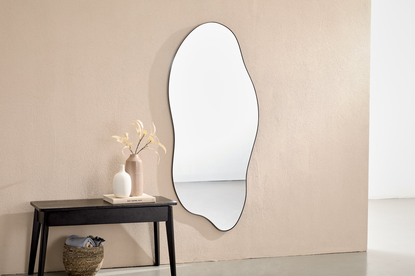 Organisch geformter Wandspiegel mit schwarzem Rahmen über einer schwarzen Konsole mit Deko-Vasen vor beiger Wand, frontal aufgenommen.