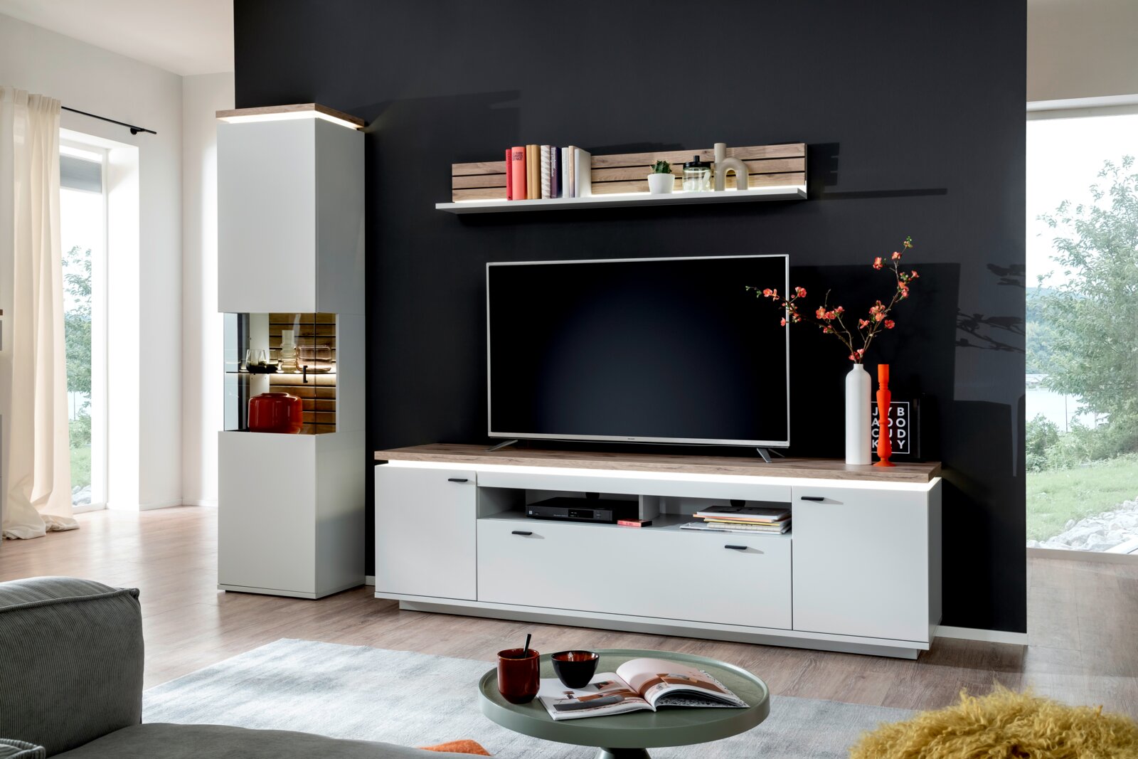 Moderne woonkamerwand in wit met afdekplaten in houtlook, tv-lowboard, wandrek en hoge vitrinekast met glazen vak voor een donkere accentmuur, van voren gefotografeerd vanuit een licht schuin perspectief van links
