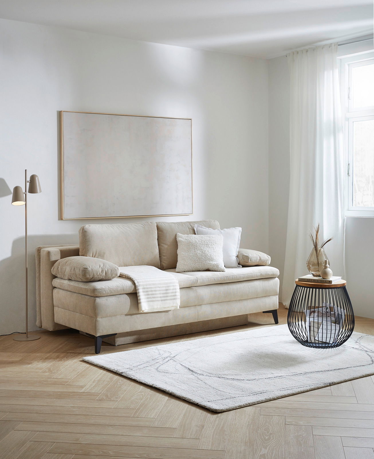 Beige Querschläfer-Sofa mit Hubbeschlag in einem modernen Wohnzimmer, seitliche Perspektive. Dekoriert mit Kissen und einer Decke, daneben ein runder Beistelltisch und eine Stehlampe.