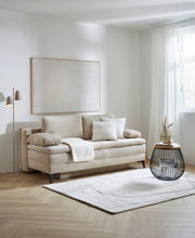 Beige Querschläfer-Sofa mit Hubbeschlag in einem modernen Wohnzimmer, seitliche Perspektive. Dekoriert mit Kissen und einer Decke, daneben ein runder Beistelltisch und eine Stehlampe.