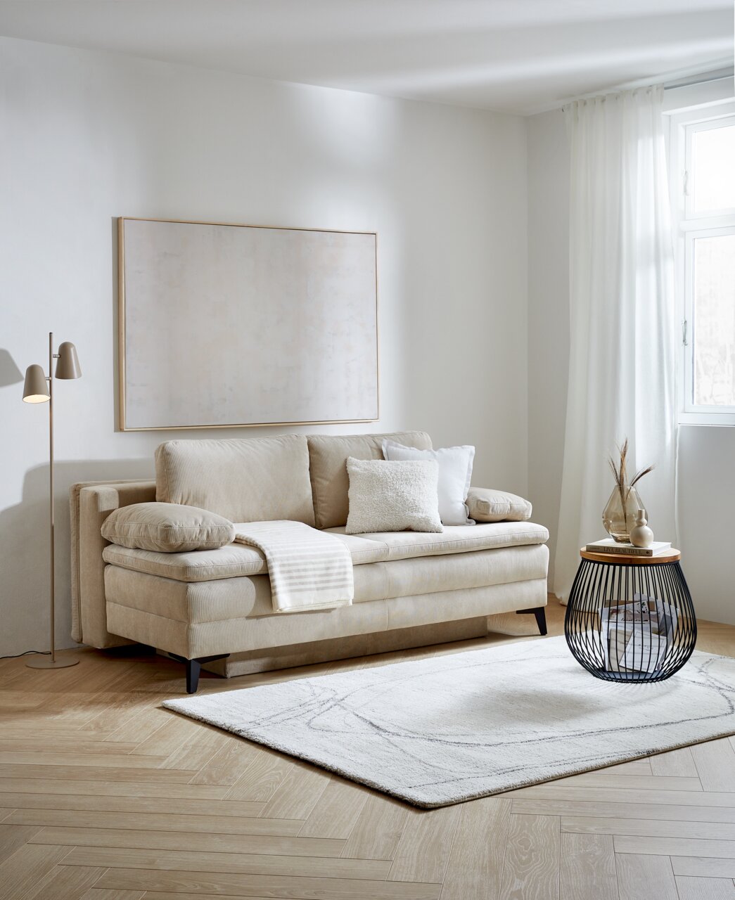 Beige Querschläfer-Sofa mit Hubbeschlag in einem modernen Wohnzimmer, seitliche Perspektive. Dekoriert mit Kissen und einer Decke, daneben ein runder Beistelltisch und eine Stehlampe.