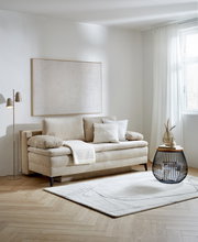 Beige Querschläfer-Sofa mit Hubbeschlag in einem modernen Wohnzimmer, seitliche Perspektive. Dekoriert mit Kissen und einer Decke, daneben ein runder Beistelltisch und eine Stehlampe.