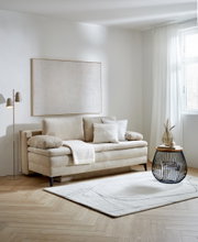 Beige Querschläfer-Sofa mit Hubbeschlag in einem modernen Wohnzimmer, seitliche Perspektive. Dekoriert mit Kissen und einer Decke, daneben ein runder Beistelltisch und eine Stehlampe.