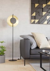 Moderne Stehleuchte TOCINA in einem Wohnzimmer, seitliche Perspektive, mit schwarzem Ständer und goldener, runder Leuchte neben einem grauen Sofa.