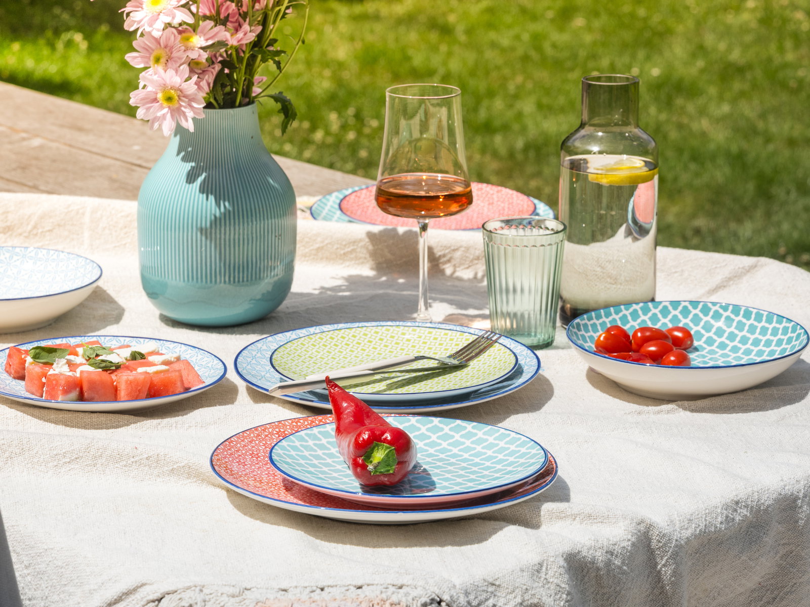 Bunte Geschirrkollektion auf einem Tisch im Freien, bestehend aus Tellern mit geometrischen Mustern, einem Weinglas, einer Vase mit Blumen und einer Karaffe mit Wasser. Perspektive von oben.