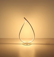 Moderne LED-Tischleuchte in tropfenförmigem, gebogenem Design aus silberfarbenem Metall mit warmweißem Licht auf runder Basis, frontal auf Augenhöhe fotografiert