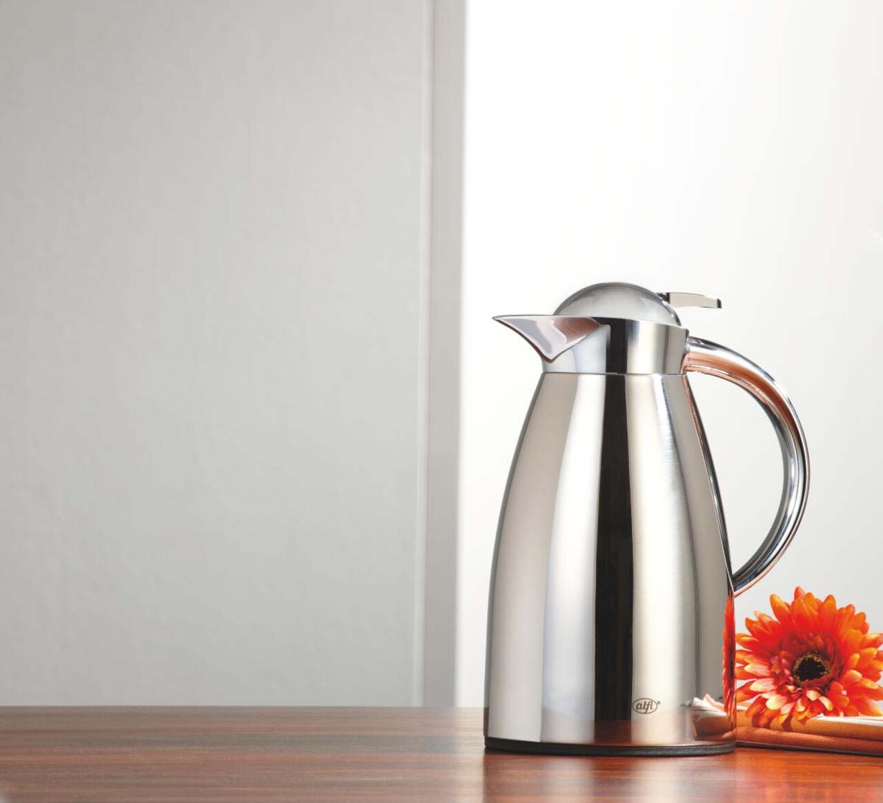 Edelstahl-Isolierkanne Signo, poliert, seitliche Perspektive, auf einem Tisch mit einer orangefarbenen Blume.