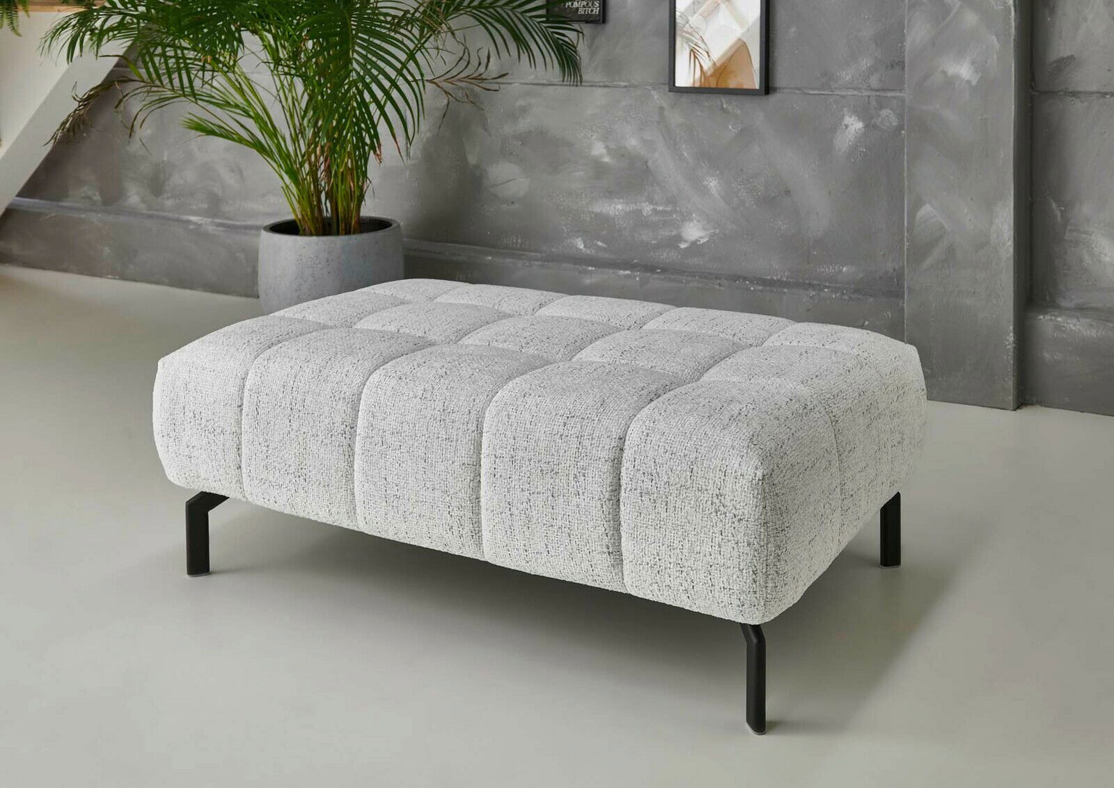 Beldomo Style XXL-Hocker VEGAS XXL-Hocker in modernem Design, seitliche Perspektive, mit grauem Stoffbezug und schwarzen Metallbeinen, vor einer grauen Wand mit Pflanze im Hintergrund.