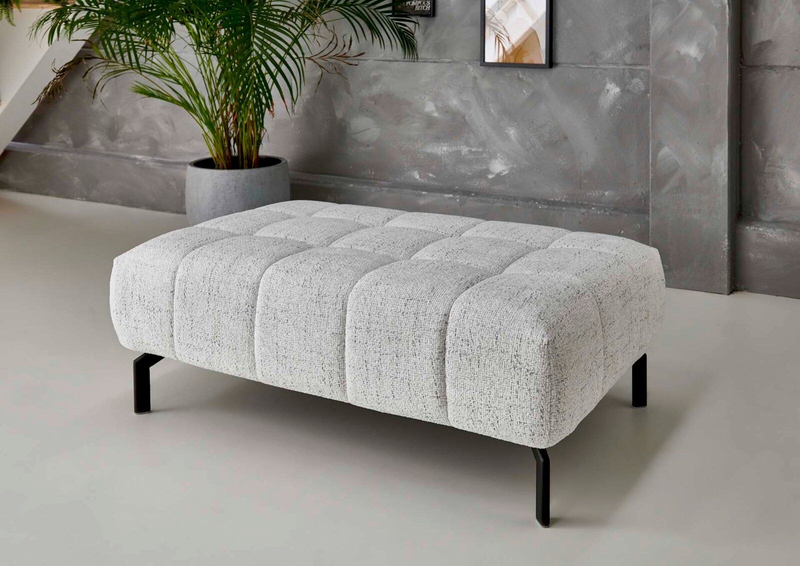 Beldomo Style XXL-Hocker VEGAS XXL-Hocker in modernem Design, seitliche Perspektive, mit grauem Stoffbezug und schwarzen Metallbeinen, vor einer grauen Wand mit Pflanze im Hintergrund.