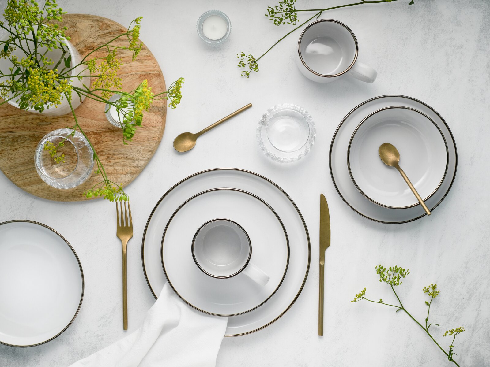 Elegantes weißes Geschirrset mit feinem Goldrand, kombiniert mit goldfarbenem Besteck, Gläsern und Blumen auf hellem Tisch – Draufsicht (Flatlay).