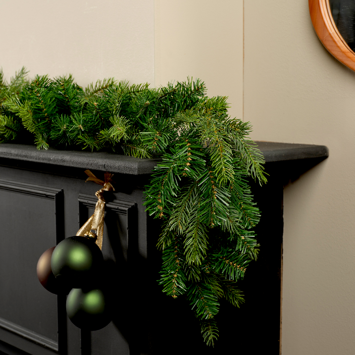 Perspektive: Seitliche Ansicht einer grünen Grandis Girlande, die dekorativ auf einem schwarzen Kaminsims liegt, mit hängenden Weihnachtskugeln.
