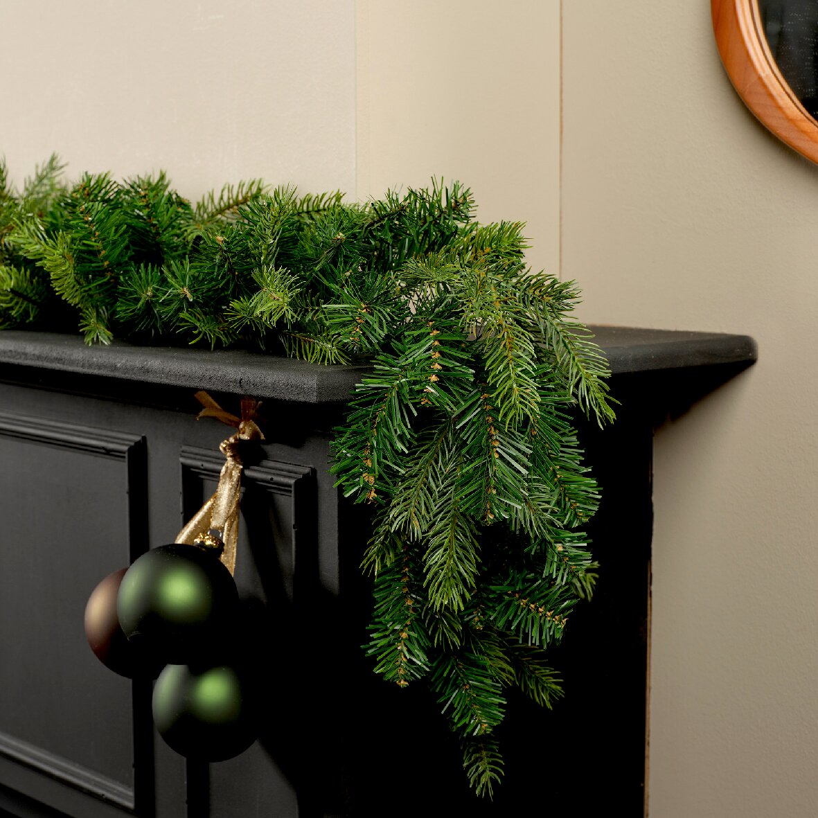 Everlands Weihnachts-Girlande 270cm Perspektive: Seitliche Ansicht einer grünen Grandis Girlande, die dekorativ auf einem schwarzen Kaminsims liegt, mit hängenden Weihnachtskugeln.