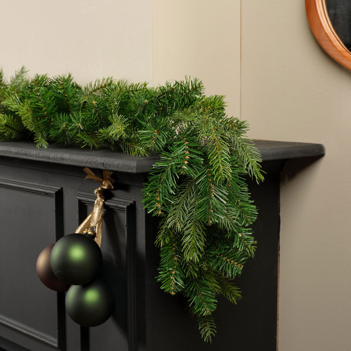 Perspektive: Seitliche Ansicht einer grünen Grandis Girlande, die dekorativ auf einem schwarzen Kaminsims liegt, mit hängenden Weihnachtskugeln.