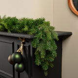 Perspektive: Seitliche Ansicht einer grünen Grandis Girlande, die dekorativ auf einem schwarzen Kaminsims liegt, mit hängenden Weihnachtskugeln.