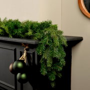 Perspektive: Seitliche Ansicht einer grünen Grandis Girlande, die dekorativ auf einem schwarzen Kaminsims liegt, mit hängenden Weihnachtskugeln.