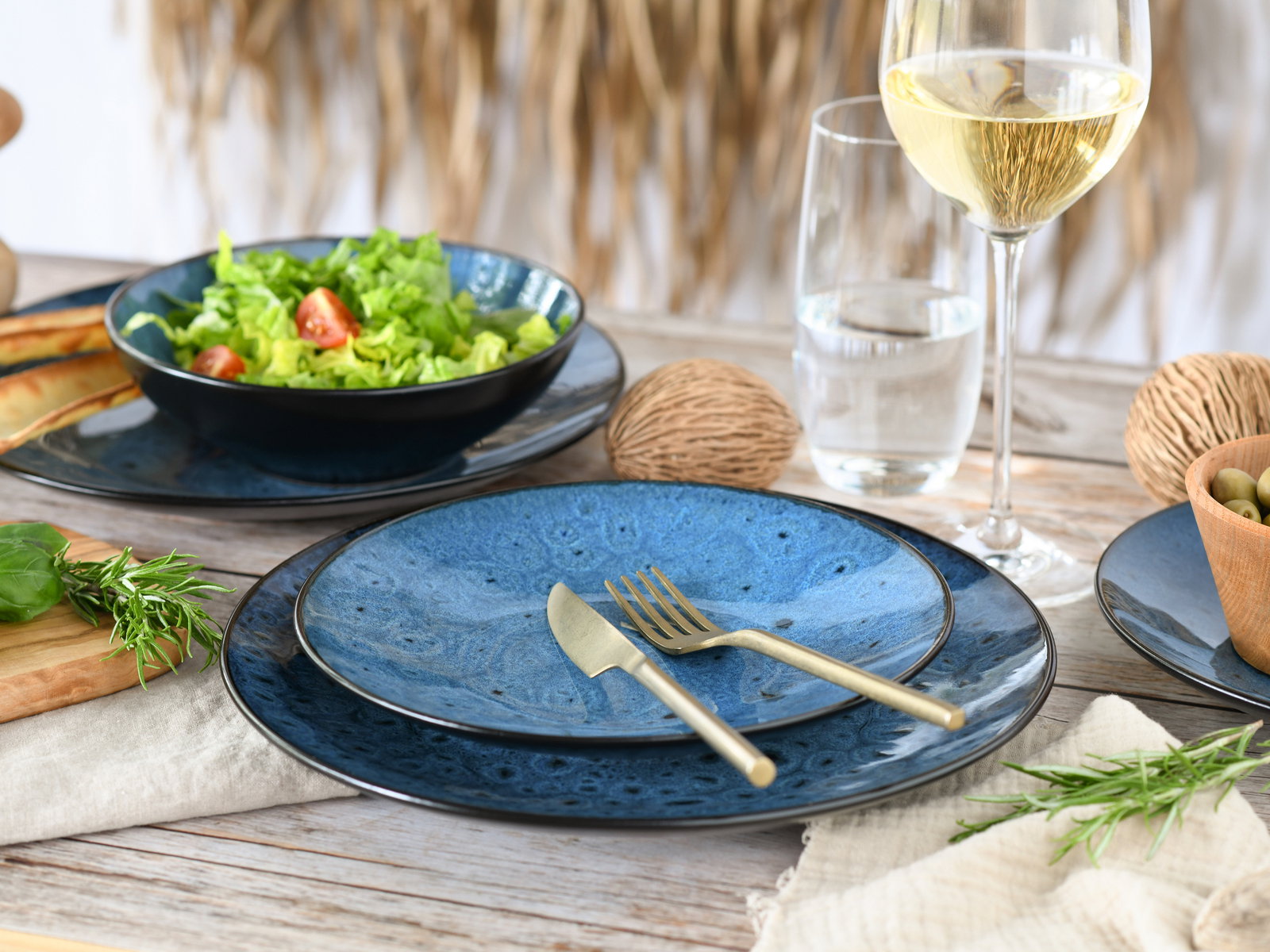 Elegantes Tellerset in Blau mit goldfarbenem Besteck, präsentiert auf einem gedeckten Tisch mit Salat, Wein und Oliven, aus der Vogelperspektive fotografiert.