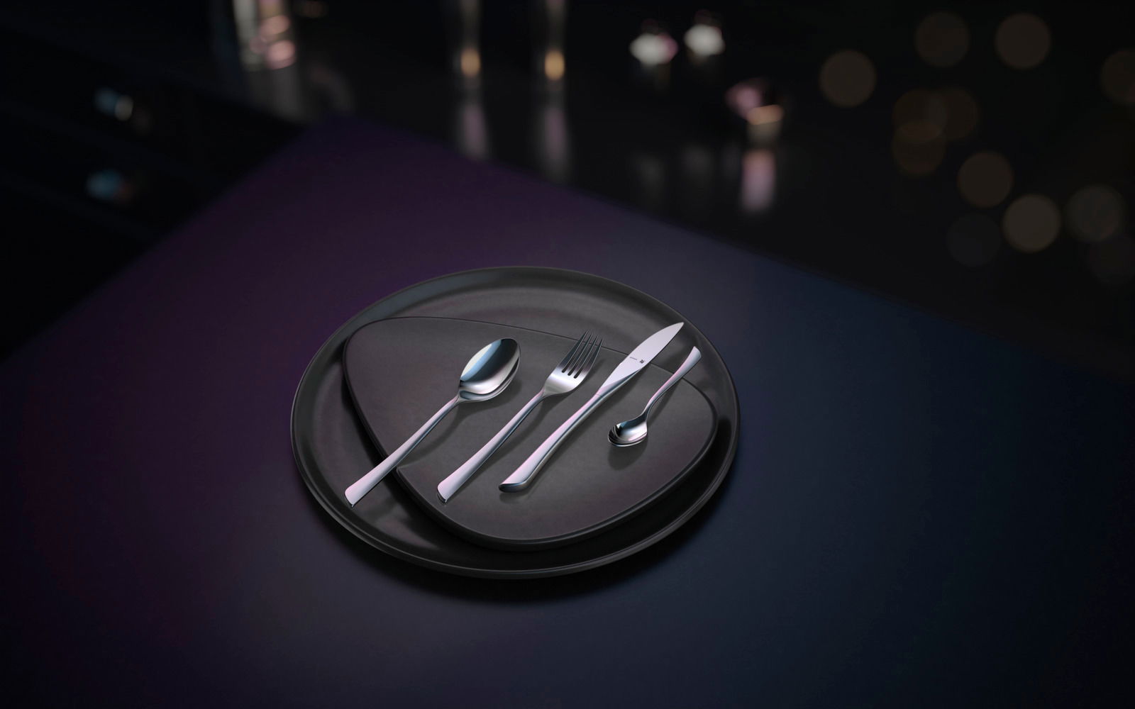 Elegantes Besteckset Virginia, bestehend aus Messer, Gabel, Löffel und Teelöffel, auf einem modernen schwarzen Teller arrangiert. Ansicht von oben.