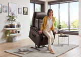 Frau sitzt in einem braunen Relax-Sessel in einem modernen Wohnzimmer mit großen Fenstern, seitliche Perspektive.
