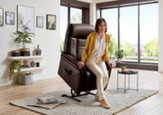 Een vrouw zit in een bruine relaxfauteuil in een moderne woonkamer met grote ramen, gezien vanuit een zijdelingse hoek.