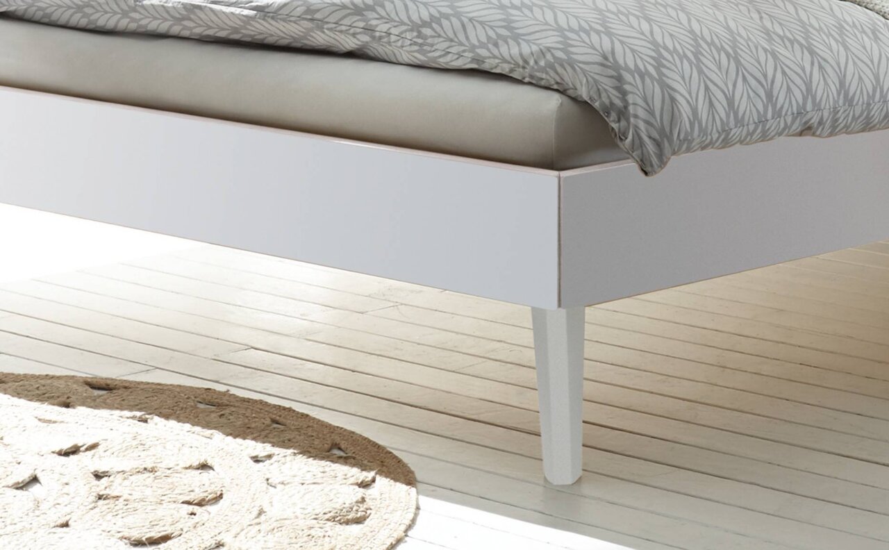 Nahaufnahme eines weißen Bettgestells mit schrägen Beinen, von der Seite betrachtet.