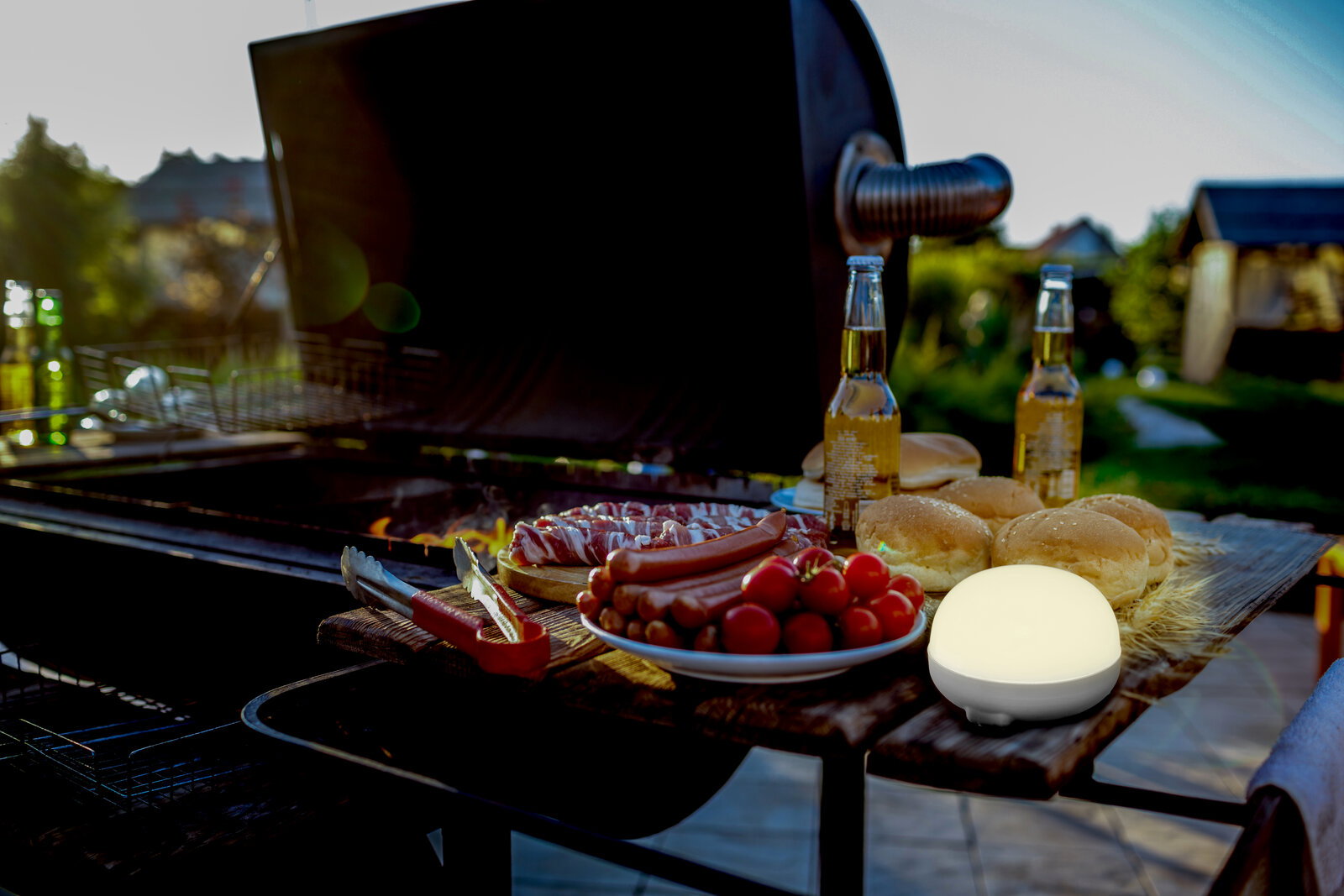 Leuchtende, runde LED-Außenlampe auf Holztisch neben Grill mit Würstchen, Tomaten, Brötchen und Bierflaschen im Garten bei Sonnenuntergang, aufgenommen aus seitlicher Nahperspektive.