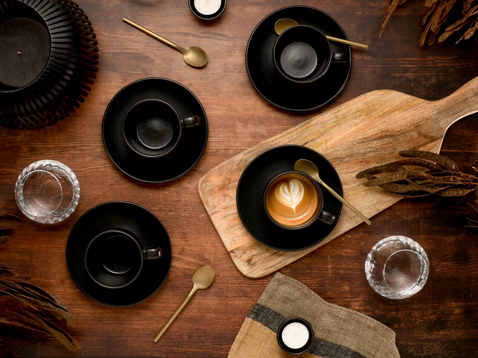 Draufsicht auf ein stilvolles Kaffee-Arrangement: schwarze Espressotassen mit Untertassen, goldene Löffel und ein Cappuccino mit Latte-Art auf einem Holzbrett auf rustikalem Holztisch.