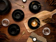 Draufsicht auf ein stilvolles Kaffee-Arrangement: schwarze Espressotassen mit Untertassen, goldene Löffel und ein Cappuccino mit Latte-Art auf einem Holzbrett auf rustikalem Holztisch.