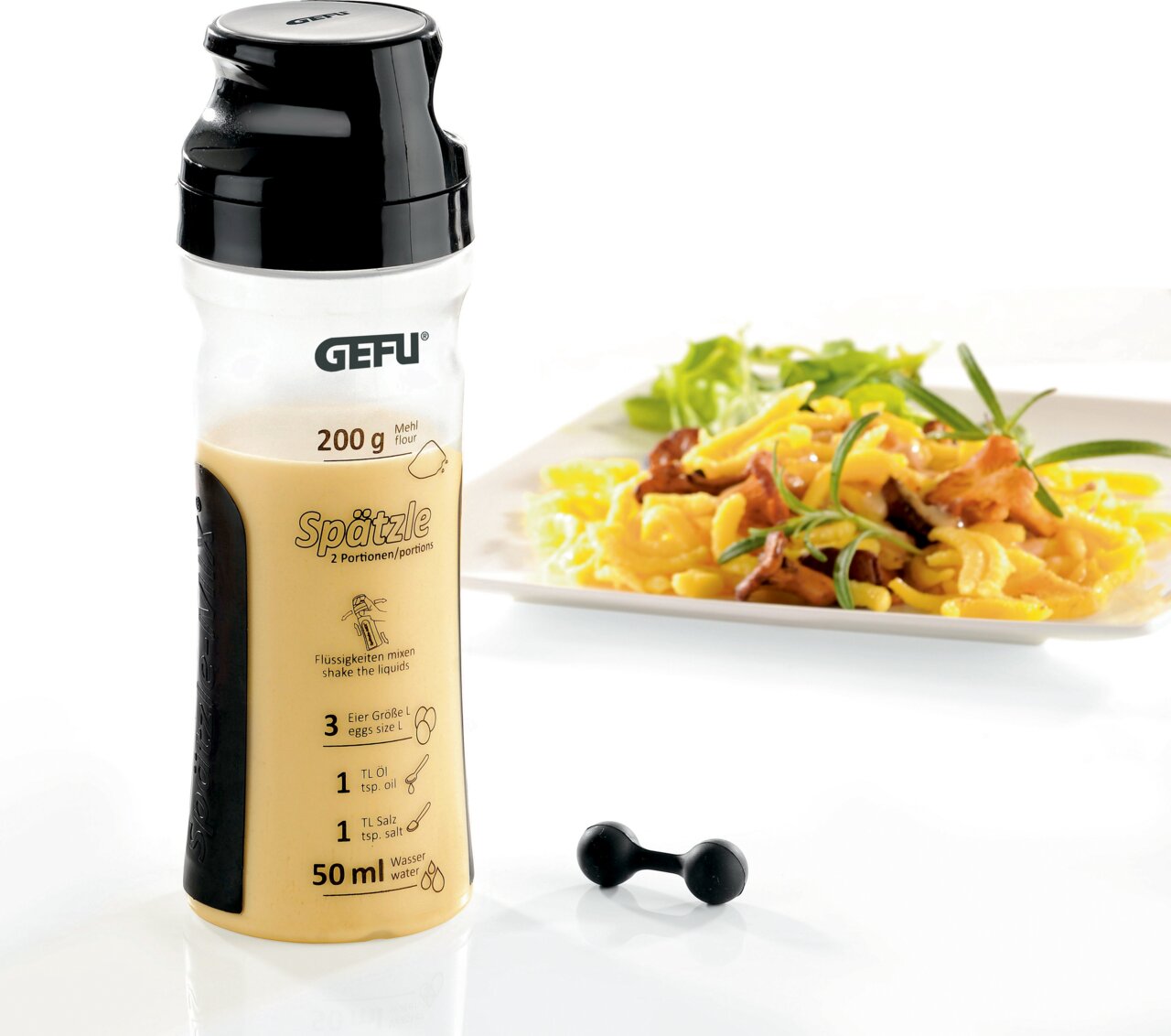 Spätzle-Mix in einer transparenten Flasche mit schwarzem Deckel, daneben ein Teller mit zubereiteten Spätzle und Kräutern, aus der Perspektive von vorne.