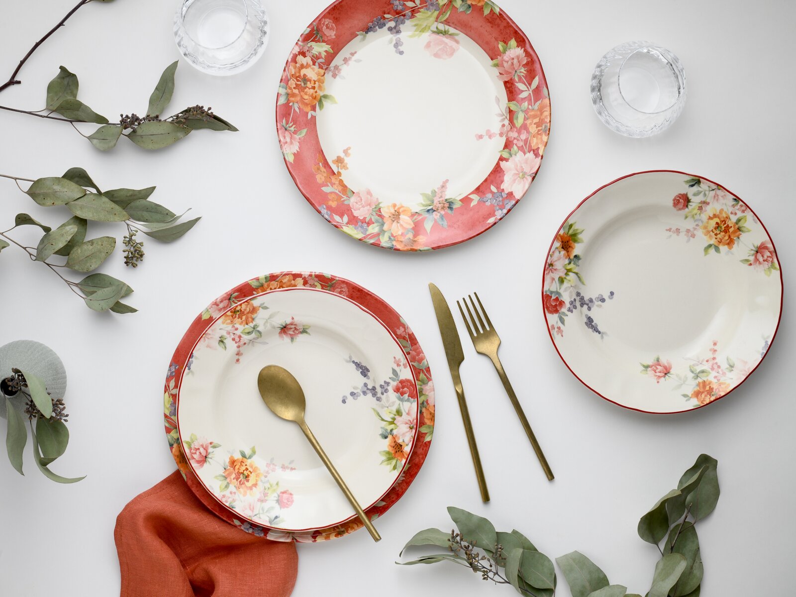 Florales Tafelservice mit roten Tellerrändern und Blütenmuster, dazu goldfarbenes Besteck und Wassergläser auf weißem Hintergrund, Draufsicht (Top-Down-Perspektive)