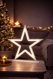 Een stralende witte led-ster op een voetstuk op een houten tafel in de woonkamer, een warme kerstsfeer met een kaars en een lichtslinger op de achtergrond, vooraanzicht