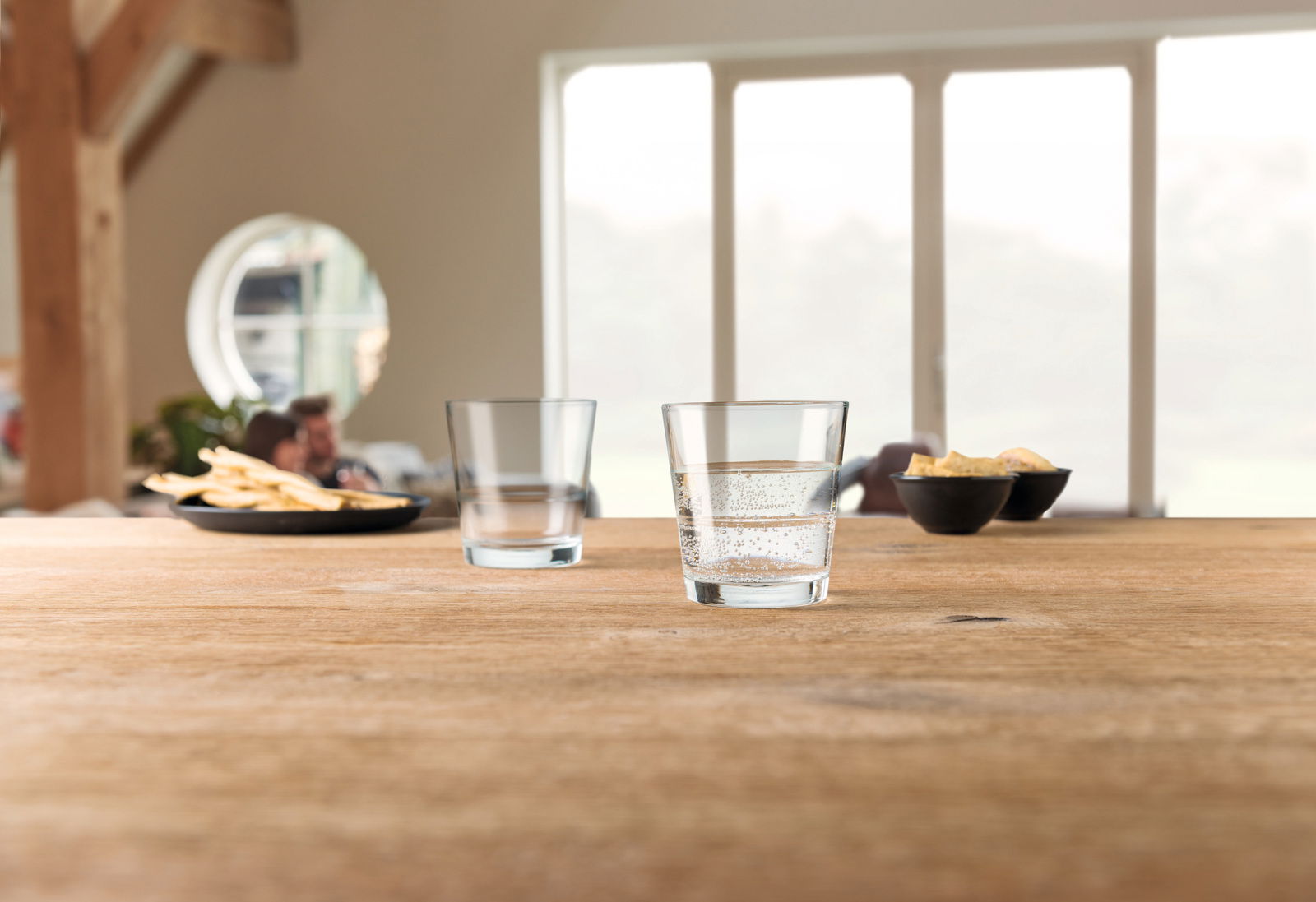 Zwei Trinkgläser Event 280ml auf einem Holztisch, eines mit sprudelndem Wasser gefüllt, im Hintergrund unscharfe Snacks und Personen, seitliche Perspektive.