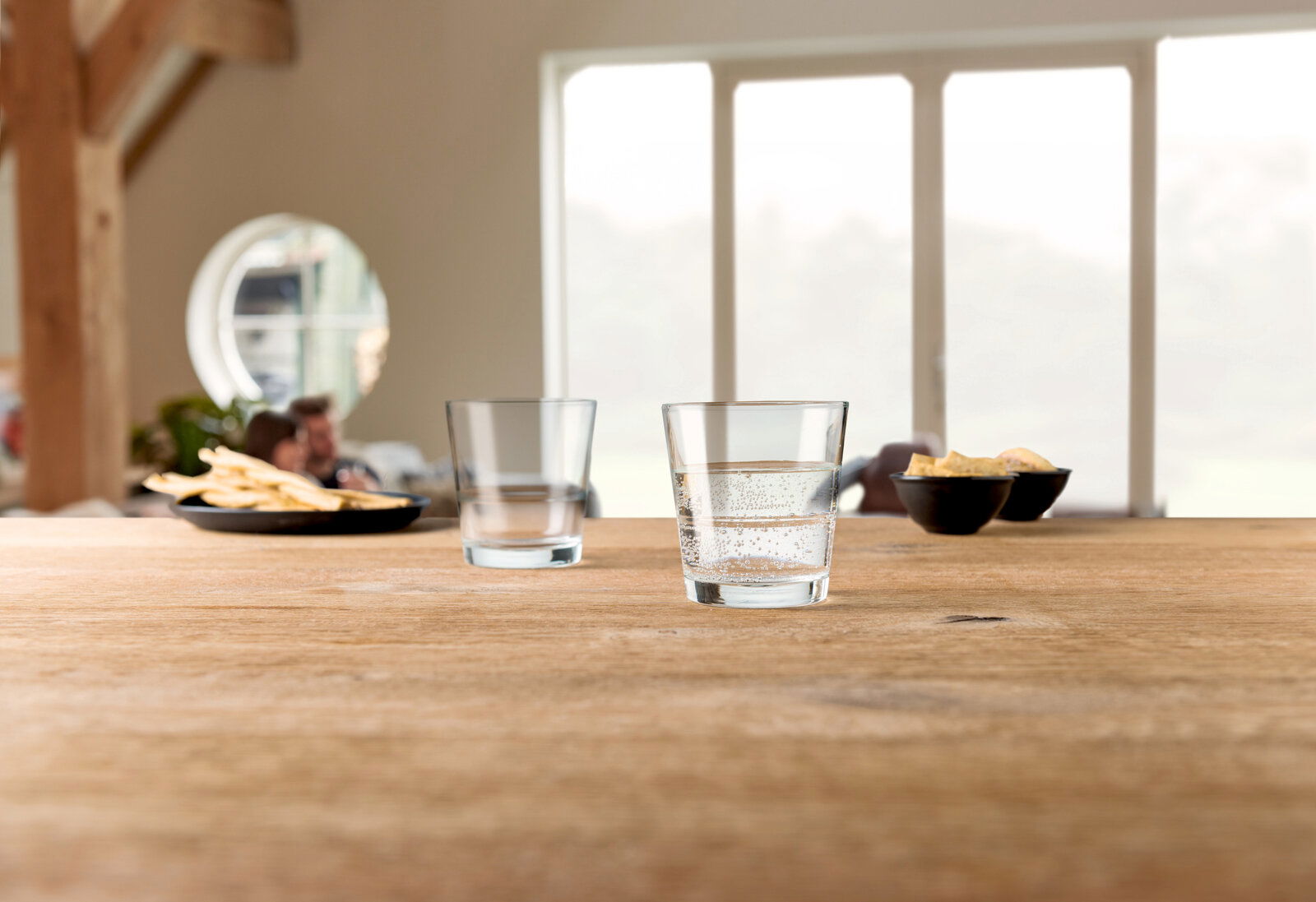 Zwei Trinkgläser Event 280ml auf einem Holztisch, eines mit sprudelndem Wasser gefüllt, im Hintergrund unscharfe Snacks und Personen, seitliche Perspektive.