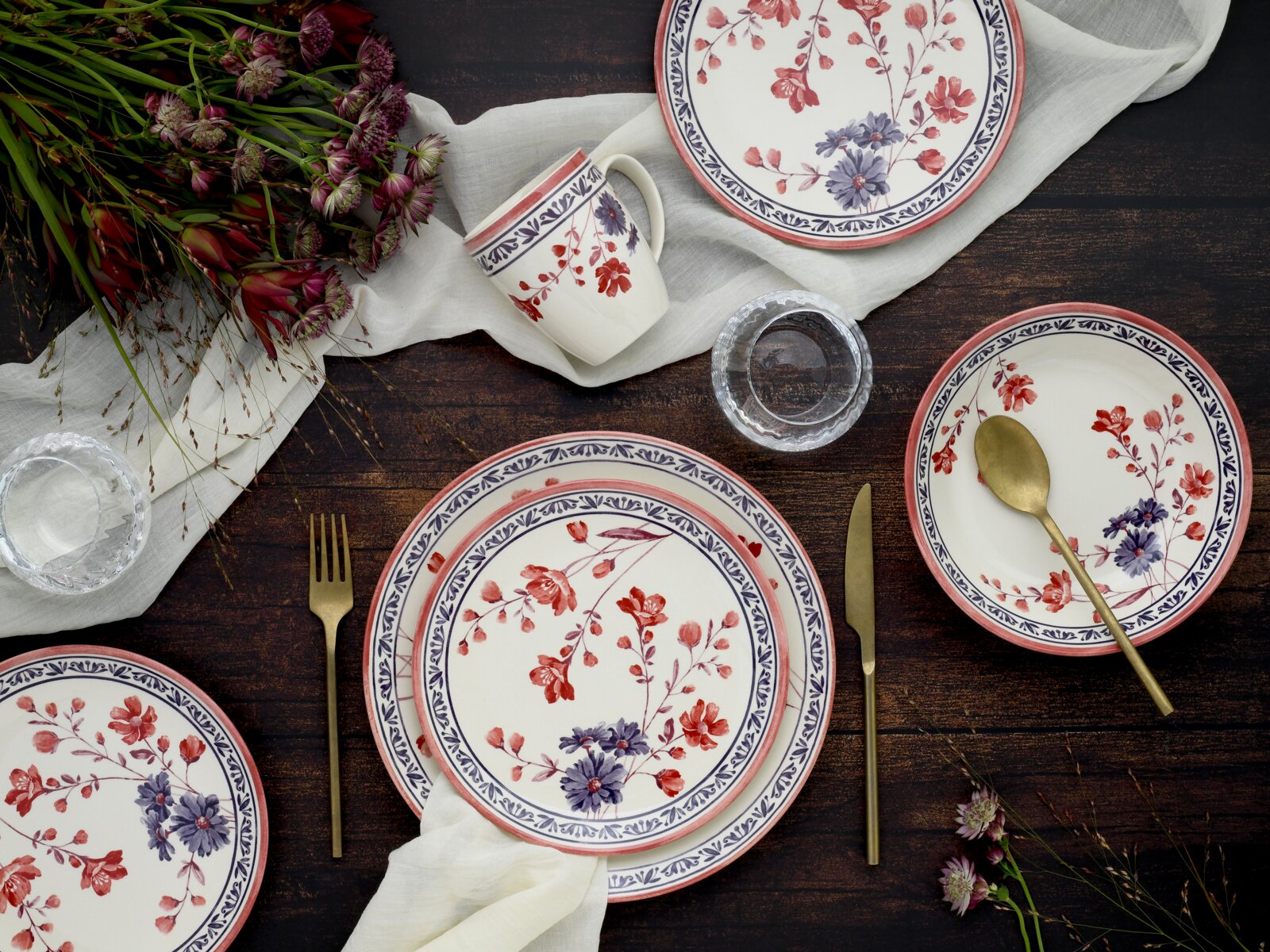 Von oben fotografiertes Tisch-Setting mit floral gemustertem Geschirr: Teller, Schale und Tasse in Weiß mit roten und blauen Blumen sowie blauem Randdekor, dazu goldfarbenes Besteck, Wassergläser, weißes Tuch und Blumen auf dunklem Holztisch.