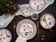 Von oben fotografiertes Tisch-Setting mit floral gemustertem Geschirr: Teller, Schale und Tasse in Weiß mit roten und blauen Blumen sowie blauem Randdekor, dazu goldfarbenes Besteck, Wassergläser, weißes Tuch und Blumen auf dunklem Holztisch.