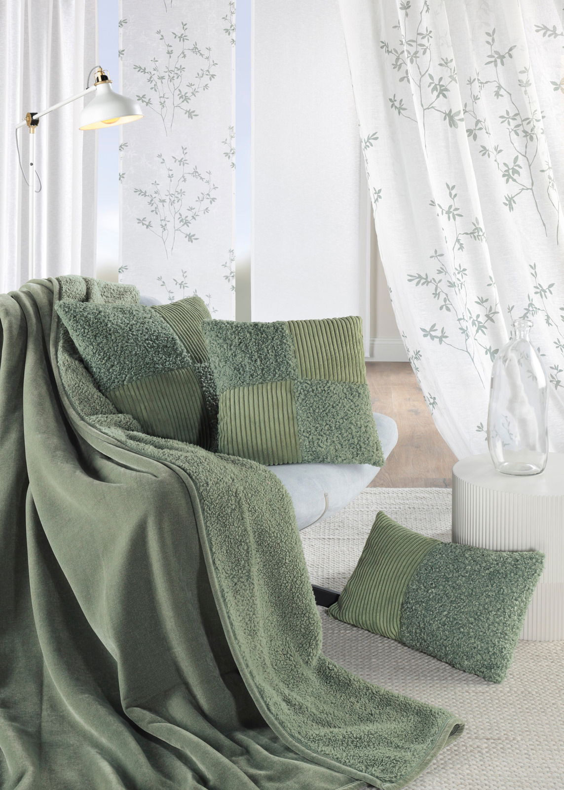 Grüne Kissenhüllen und Decke auf einem Sofa, umgeben von weißen Vorhängen mit Pflanzenmuster, aus der Perspektive von vorne.