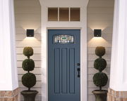 Moderne schwarze Akku-Wandleuchte an einer Hausfassade neben einer blauen Eingangstür, von vorne betrachtet.