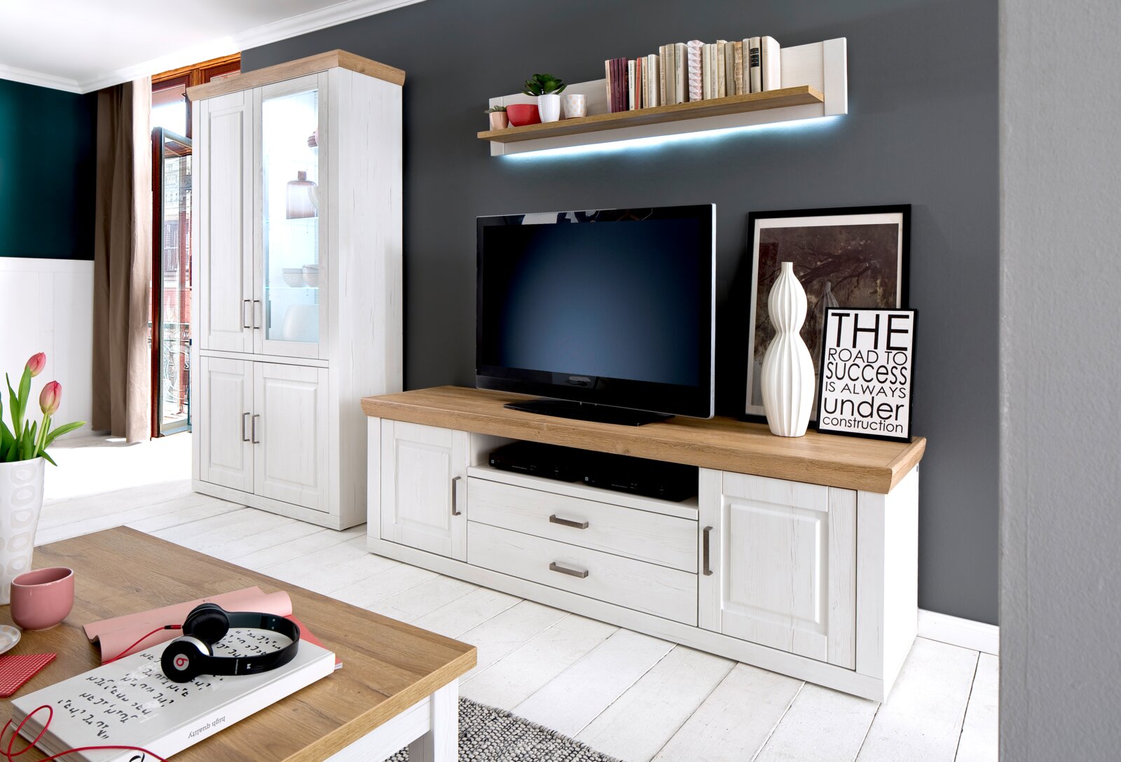 Landscape Wandcombinatie BREGENZ Woonkamermeubelset in landhuisstijl met een witte vitrinekast en een tv-lowboard met eikenhoutlook, een aan de muur bevestigde plank met ledverlichting en een televisie; perspectief: vooraanzicht met een lichte zijaanzicht.