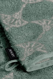 Nahaufnahme eines grünen Waschhandschuhs mit dekorativem Muster und einer schwarzen Schlaufe mit der Aufschrift 'JOOP!'.