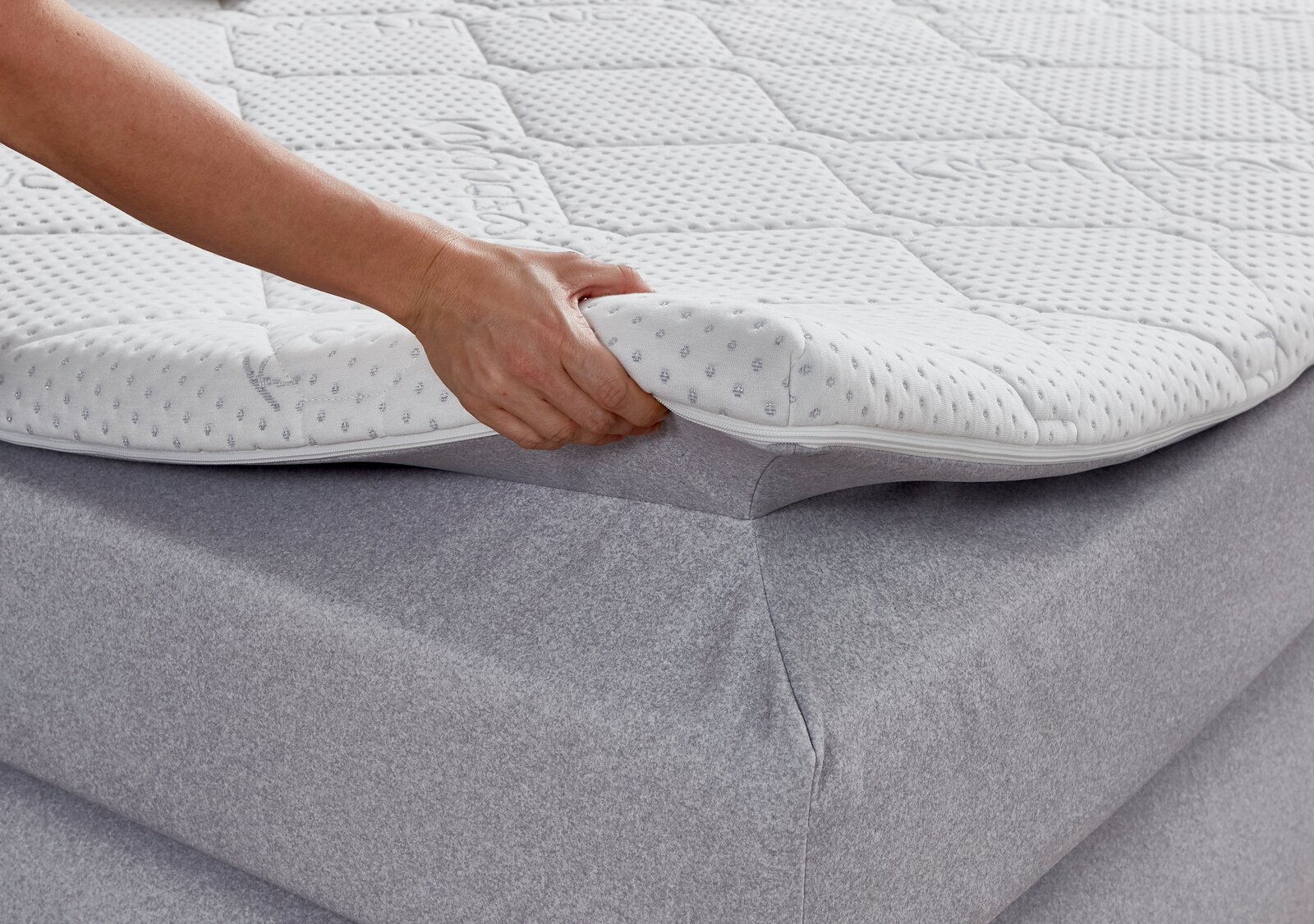 Nahaufnahme eines Boxspringbetts mit einer Hand, die den Matratzentopper anhebt, um die weiche Polsterung und den Bezug zu zeigen.